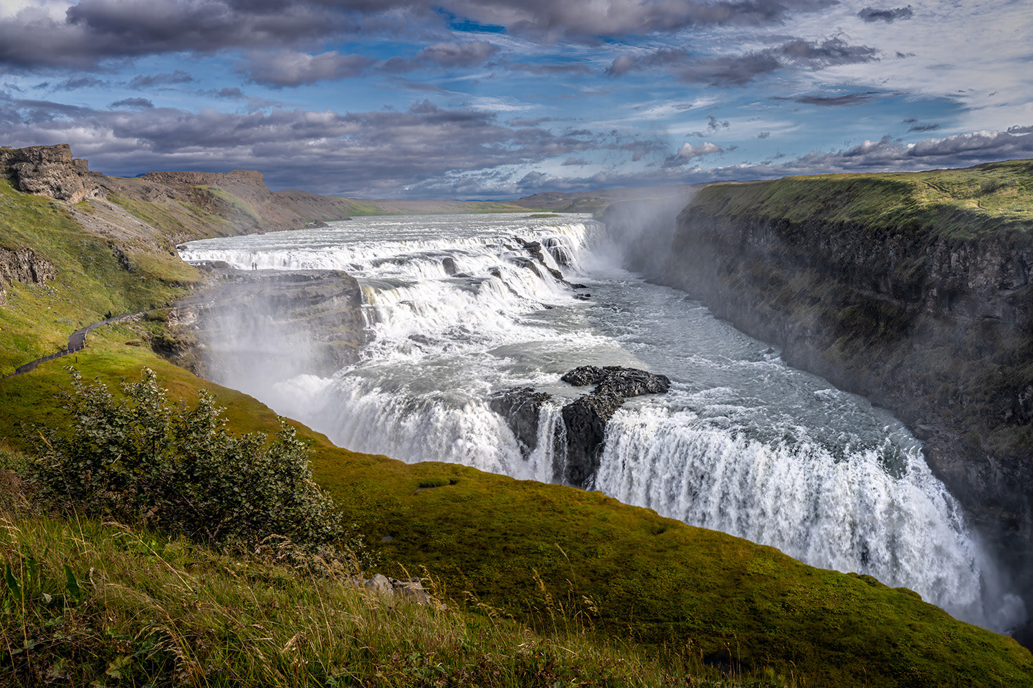 Prachtige weergave van de Gullfoss Waterval in IJsland, omringd door groene landschappen onder een wolkenlucht.