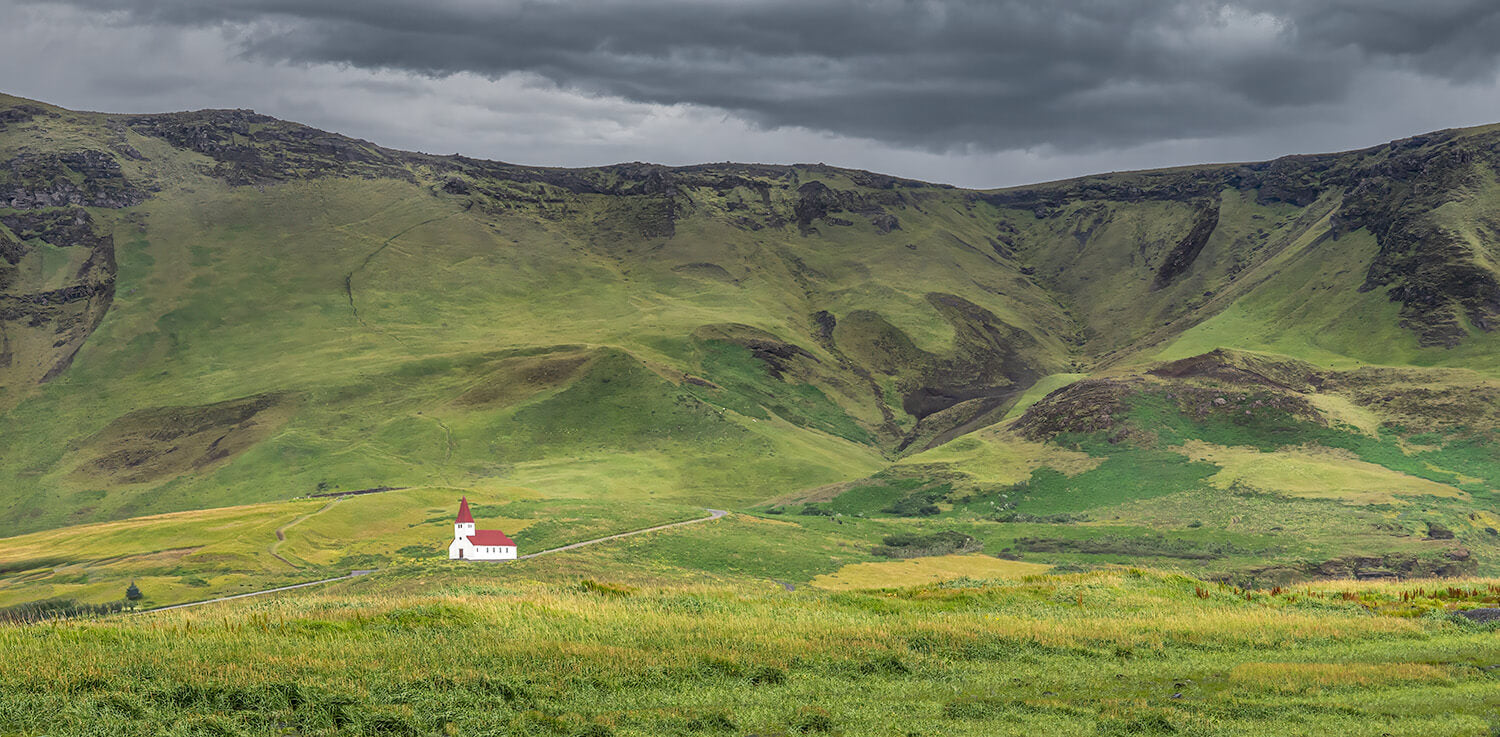 Een wit kerkje met rood dak omgeven door groene heuvels en een dramatische lucht in IJsland.