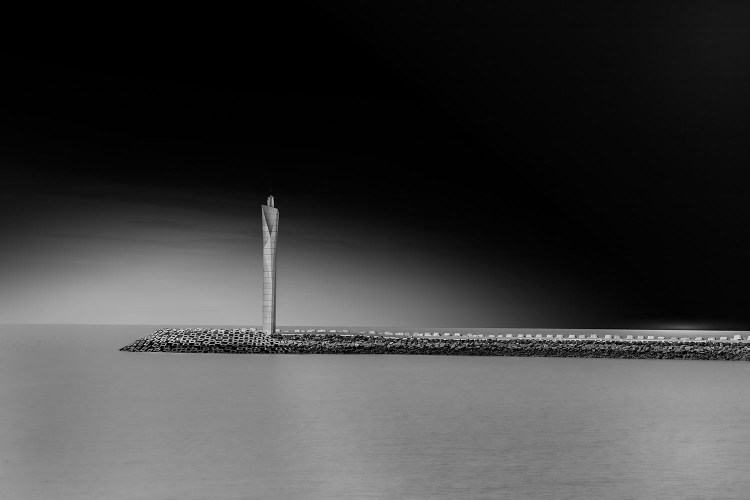 Zwart-wit foto van de moderne vuurtoren in Oostende, staande op de pier, met een dramatische, donkere lucht op de achtergrond.
