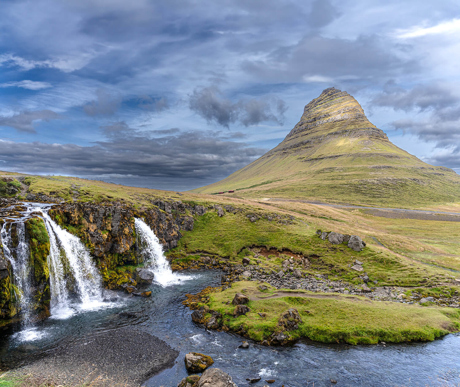 Het iconische Kirkjufell-gebergte met een waterval op de voorgrond, onder een dramatische bewolkte lucht in IJsland.