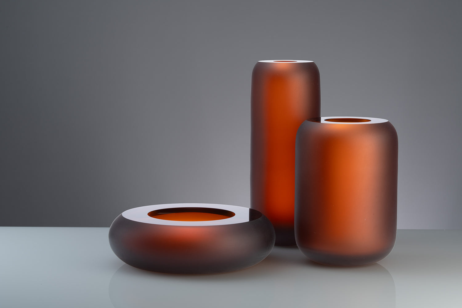 Glazen kunstobjecten uit de "Termoska Collection" door Anna Torfs, in warme oranjerode tinten tegen een minimalistische achtergrond.